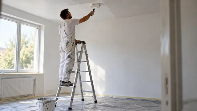 Zugrav pe o scară din aluminiu deschisă în formă de A, care vopsește tavanul unei camere goale cu trafaletul