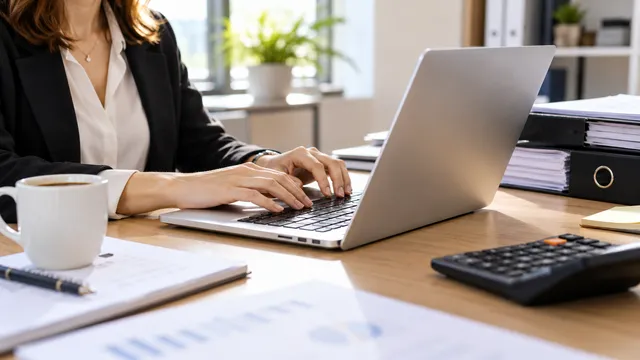 O contabilă lucrează la un laptop subțire argintiu pe un birou, lângă dosare și un calculator de birou