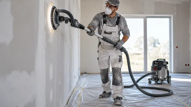 Zugrav cu mască de praf care șlefuiește un perete de gips-carton cu un șlefuitor telescopic tip girafă cu lumină LED, legat la un aspirator